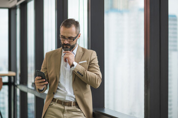 Young handsome business bearded man standing post near window using smartphone in office work place.
