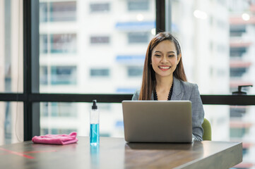 Young asian businesswoman using laptop sitting in modern office working with social distancing alcohol gel on desk.