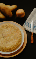 Homemade Spanish potato omelette and some onions, some potatoes, a napkin and a knife in the background