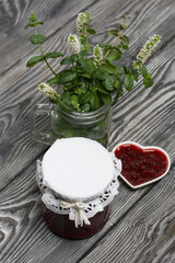 Fototapeta na wymiar Raspberry jam in jars and a saucer. On pine planks painted black and white. Nearby flowering branches of mint in a jar.