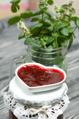 Raspberry jam in jars and a saucer. On pine planks painted black and white. Nearby flowering branches of mint in a jar.