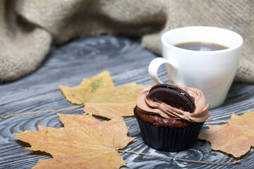 Chocolate cupcakes with cream cheese cream. Nearby is a cup of coffee and dried maple leaves. On pine planks painted black and white.