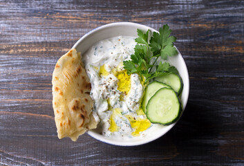Traditional greek yogurt tzatziki sauce with cucumbers and herbs on wooden background, top view	