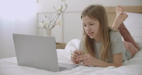 schoolgirl is communicating with teacher by video call on laptop, lying on bed in home, online lessons for secondary schoolers, distant education - Powered by Adobe