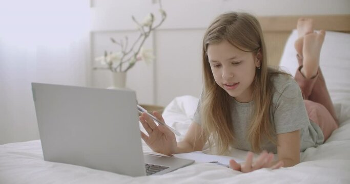 teen girl is lying on bed with laptop and exercise book and talking, preparing to doing homework after school