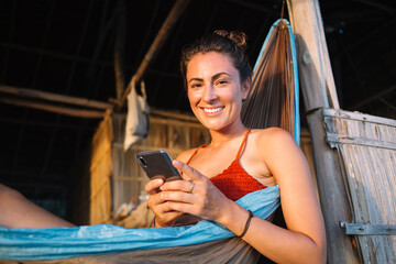 Portrait of cheerful female user with cute smile on face resting in swing hammock enjoying digital nomad lifestyle, happy woman with modern smartphone gadget for blogging recreating during vacations