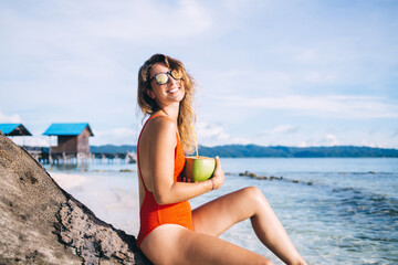 Young woman sitting on dead tree with cocktail in coconut