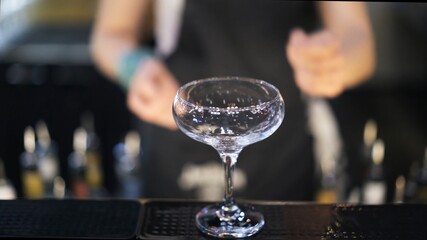 Empty cocktail glass, close up, on blurry background of bartender at bar. Cocktail glass focused shot and barman inside on blurred background, making cocktail concept