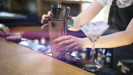 Metal cups on bar counter, bartender holding cups to mix cocktail. Cups and glass with ice on bar counter on background of barman hands, lens flare