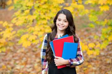 Fototapeta premium concept of knowledge day. kid in autumn park. happy kid with books in autumn leaves. small girl wear uniform. back to school. modern education. child with notebooks in fall forest