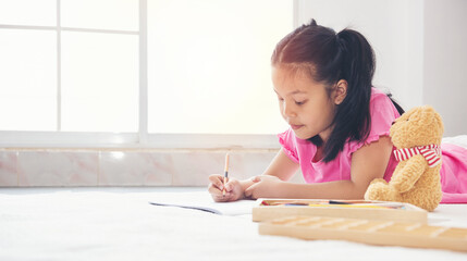 Little asian girl in pink shirt practicing writing laying on the floor in her room with bear doll. child girl writing letter near window.