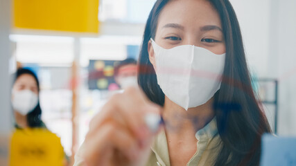 Asia young businesspeople discussing business brainstorming meeting working together sharing data and writing glass wall with medical face mask back at work in office. Life and work after coronavirus.