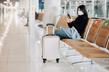 Asian woman tourist wearing face mask sitting on social distancing chair with luggage during...