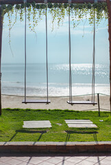 Two wooden swings next to each other are tied to the bar on a sunny morning. The blurred background is a blue ocean with glitter. The top edge of the image has ivy and the lower has green grass.