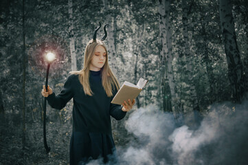 A young girl with long hair in a witch costume, with a staff in her hands, conjures a book. Halloween, selective focus, smoke in the woods. Tinting for an old photo with a grainy overlay.
