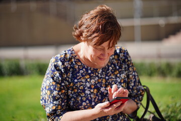 Adult woman sitting on a Park bench, looking at her mobile phone. A woman using a smartphone uses the Internet in the courtyard of a residential building.
