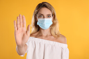 Woman in protective face mask showing stop gesture on yellow background. Prevent spreading of coronavirus