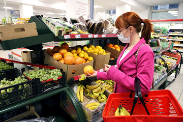 Young woman wearing a protective face mask, holding an orange in a supermarket. She stands by the shelves with vegetables and fruits and puts food in a basket.