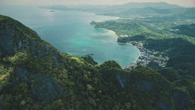 Mountainous tropical island aerial: pier town cityscape with vessels, boats on water surface. Green palm trees on mountain ranges. Houses, homes, lodges at tropic resort. Cinematic drone view