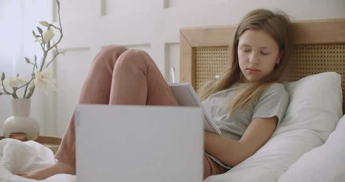 tired and bored pupil girl is lying on bed with exercise book and listening lesson on laptop, e-learning for school children