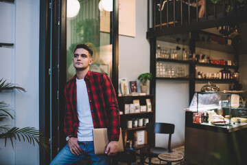 Cheerful man with laptop standing near entrance of cafe