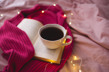 Cup of coffee with open book and knitted clothes over glowing lights in bed close up. Selective focus.