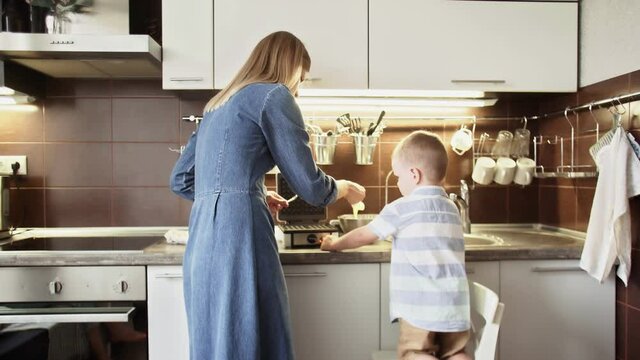 Young mother and her son are making waffles in waffle maker at home kitchen