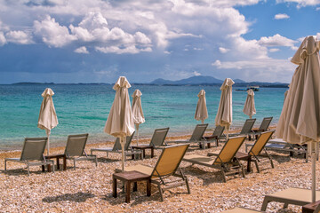 View of empty beach - white deck chairs and umbrellas near sea water, pebbles and golden sand, mountains on the horizon and dark clouds on the sky. Landscape of abandoned summer resort.