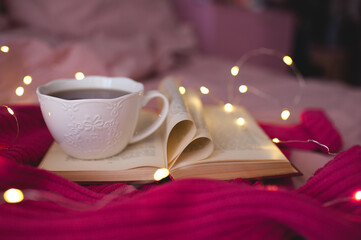 Cup of hot black tea staying on open book with heart shape folded pages and knitted pink sweater over glowing Christmas lights closeup in bed. Winter holiday season. Selective focus.