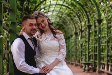 Bride and groom sit embracing in Park with an artificial vineyard in background. Newlyweds in wedding dresses on Sunny wedding day. Couple on street in amazing view. Newlyweds in love happy together