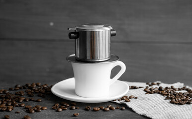 White cup of coffee with a metal filter on a top and scattered beans on a wooden table. Traditional vietnamese coffee.
