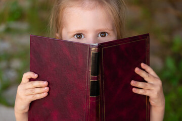 Shocked child holding a book in her hands and looking at the camera. Emotions of a schoolgirl reading a book. Knowledge and learning.