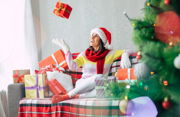 Celebration of the winter holidays. Beautiful happy brunette woman in santa hat and sweater is having fun with many gift boxes on the couch at home. Merry Christmas and happy new year