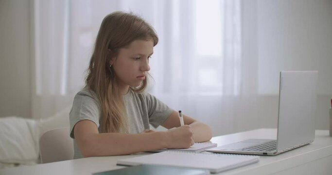 teen girl is learning distantly at home, looking at screen of laptop and writing in exercise book