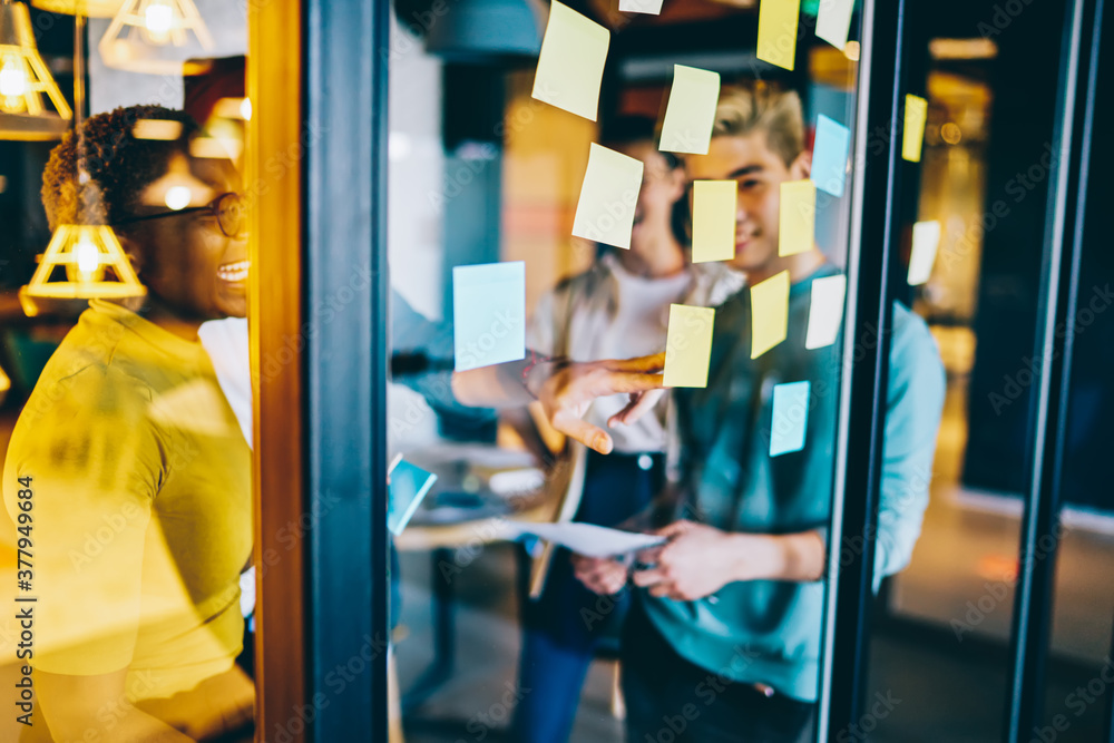Poster group of happy hipster guys making notes on colorful memo sticks at glass wall while creating intere