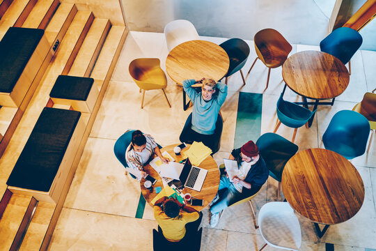 Top View Of Male And Female Students Sitting At Round Table Desktop And Collaborating On University Course Work, Group Of Skilled Hipster Guys Communicate About Business Ideas During E Learning