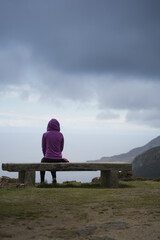 Thoughtful Hooded person is Looking at Ocean siting on a stone bench on top of a high cliff. Back...