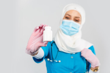 Fototapeta na wymiar friendly Muslim doctor or nurse in a hijab, mask, gloves offering a pill to the patient on a gray background