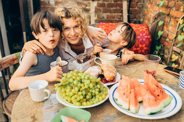 Happy middle age blond mother carring her children. Mature woman with sons eating fruits and dessert at terrace over brick wall on backyard.