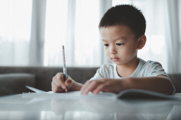 Asian Boy Doing homework with the intention. Child boy holding pencil writing, A boy drawing on white paper at the table,Elementary school and home schooling, Distance Education concept.