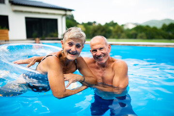 Cheerful senior couple in swimming pool outdoors in backyard, looking at camera.