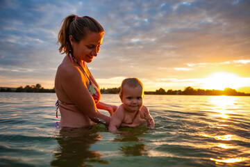 Side view of happy caucasian mother teaching swimming little lovely smiling baby in sea water. Smiling family having fun, summertime outdoors during fantastic bright orange sunset. Family concept.