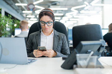 Business woman dressed in a headset is bored and uses a smartphone while sitting at a desk. Female...