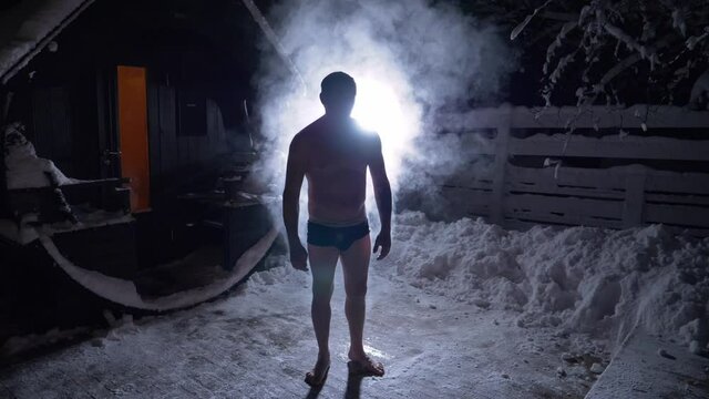 Steam rising from a backlit man, standing nude outside of a sauna, on a winter night, in Poland - Static, slow motion shot