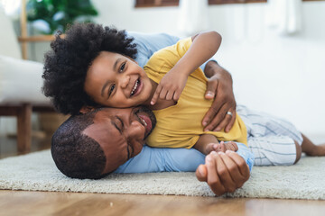 Black father in living room have a fun playing together on warm floor at home.Happy African family dad hugging, embracing his lovely child while lying on the floor.Sweet moments of fatherhood concept. - obrazy, fototapety, plakaty