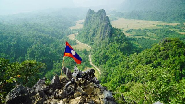 Motorcycle on top of the Nam Xay Mountain Peak with a Laos Flag Blowing and Green Forest Below