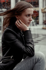 Side view portrait blode woman with windy hair wearing black leather jacket. Female model sitting on a bench and looking away