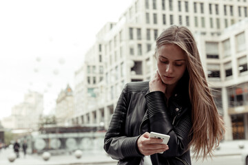 Young woman wearing black leather jacket and white shirt reading emails on a mobile phone while standing outdoors on a blurred urban background.