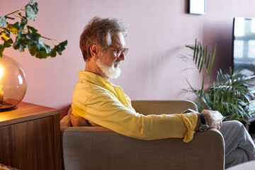 side view on senior man reading a book at home, serious male sit on chair looking at book, wearing...