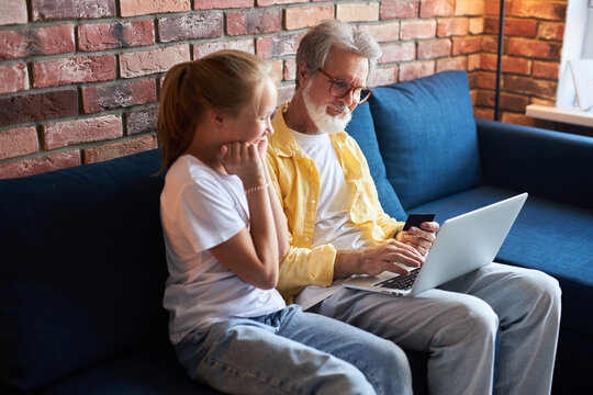 Little Girl In Anticipation Of Grandfather Making Purchase Online , Using Laptop And Credit Card. At Home